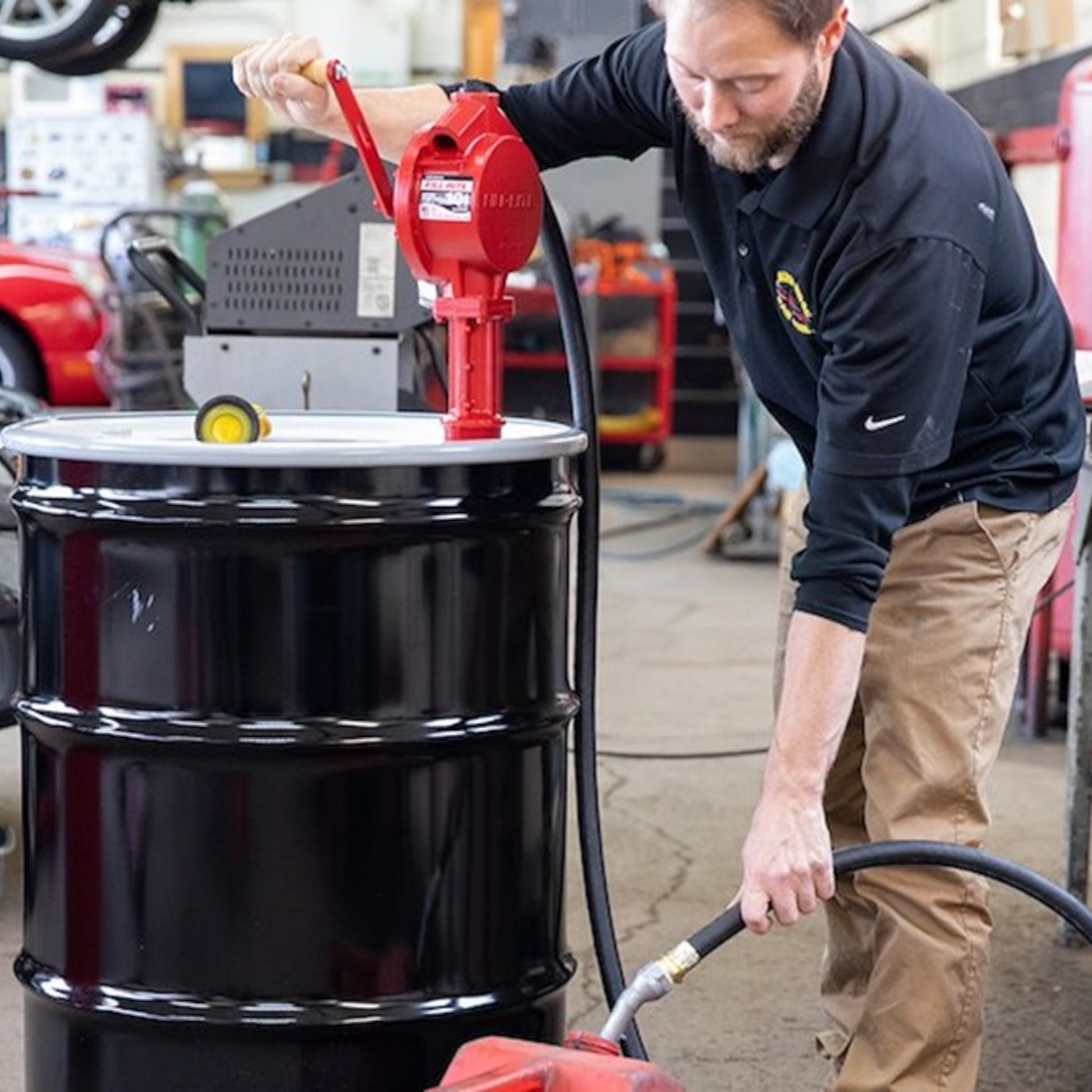 Mechanic operating Fill-Rite FR112 red rotary hand pump on drum in workshop, fueling jerry can with hose for efficient transfer