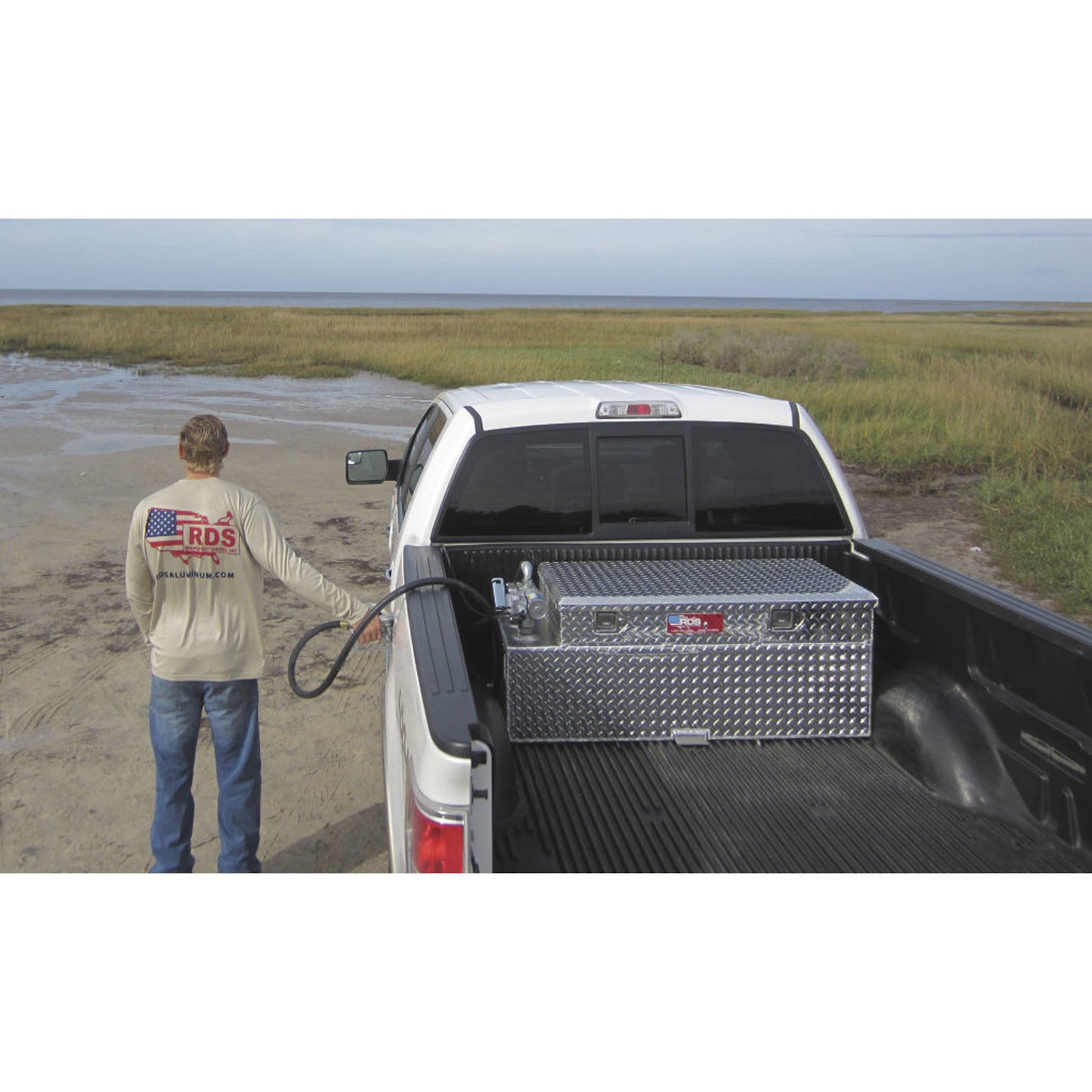 Man using RDS aluminum transfer fuel tank/toolbox combo to refuel truck in marshy terrain: portable auxiliary transfer for off-road and remote adventures.
